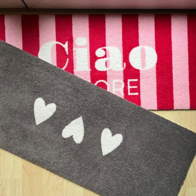 May include: Two rectangular rugs. One is gray with three white hearts. The other is red and pink striped with the word "Ciao" in white. The word "MORE" is also visible in white.