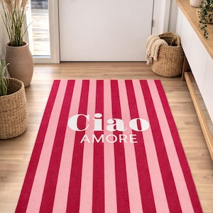 May include: A rectangular rug with alternating vertical stripes of light and dark pink. The rug features the words "Ciao AMORE" in white, centered on the rug. The rug is on a wooden floor.