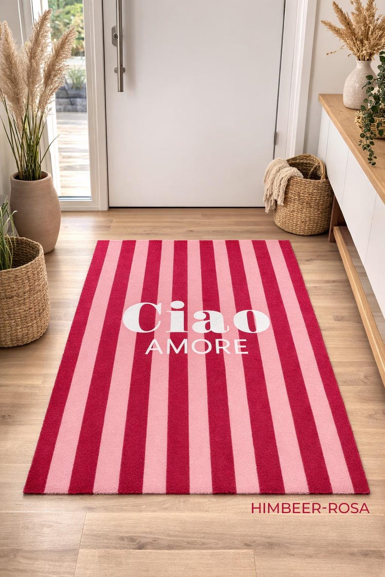 May include: A rectangular rug with alternating vertical stripes of light and dark pink. The rug features the words "Ciao AMORE" in white, centered on the rug. The rug is on a wooden floor.