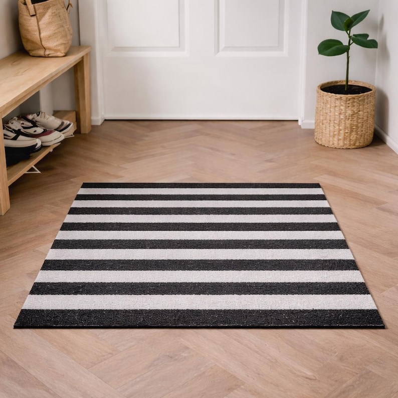May include: A rectangular black and white striped doormat. The mat is made of a textured material and is placed on a wooden floor. The stripes are horizontal and evenly spaced. The doormat is in front of a white door.