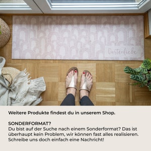May include: A rectangular, blush-colored doormat with a repeating arch pattern. The text "Gartenliebe" is visible on the mat. The mat is placed on a wooden floor, with a person's feet wearing sandals in the foreground.