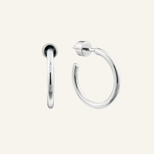 Könnte beinhalten: Zwei silberne Creolen. Jede Ohrring hat ein abgerundetes, poliertes Design. Ein Ohrring hat einen Stecker und eine kleine, flache Rückseite. Der andere Ohrring hat ein abgerundetes, kugelförmiges Ende. Die Ohrringe sind vor einem neutralen Hintergrund platziert.