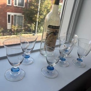 May include: A set of five clear glass goblets with blue glass accents on the stems and a tall, empty glass bottle with a label reading JOANNA and FOURNIER. The glasses feature a rounded bowl shape and a circular base, arranged on a flat surface.