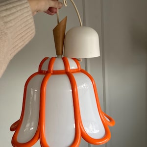 Puede incluir: Una lámpara colgante blanca y naranja con una pantalla en forma de flor. La pantalla tiene cinco pétalos con un centro blanco y bordes naranjas.