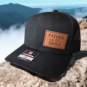Peut inclure: Casquette de camionneur noire avec un patch en cuir marron qui dit "Father of the Bride" en écriture blanche.