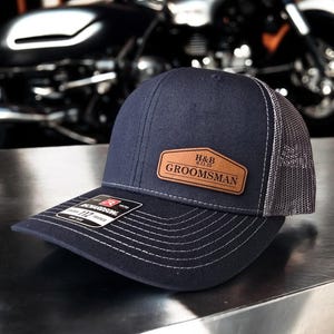 May include: A navy blue and grey trucker hat with a brown leather patch that says "H&B 6.31.23 GROOMSMAN".