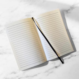 May include: A blank lined notebook with a black ribbon bookmark. The notebook is open to a blank page.