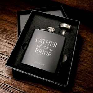 May include: Black gift box with a black metal flask engraved with "Father of the Bride" and "Andrew". The flask comes with a silver funnel.