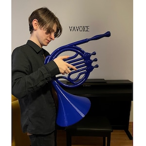 May include: A person is holding a blue, plastic French horn. The horn is large and has a bell-shaped end. The word "VAVOICE" is written on the wall behind the person.