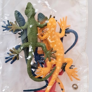 May include: A collection of plastic lizard figurines in various colors including green, orange, blue, and red, all contained within a clear plastic bag. The lizards feature textured skin, visible toes, and long tails, layered on top of one another.
