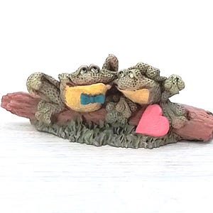 May include: A figurine of two frogs sitting on a log, with a pink heart. One frog wears a yellow vest and a blue bow tie. The frogs are green and tan, and the log is pink and brown. The base is green.