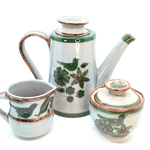 May include: A ceramic tea set featuring a teapot, creamer, and sugar bowl. The set is painted with green birds and floral designs on a cream-colored background. The rims are accented with brown and green trim.