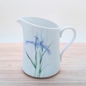 May include: White ceramic pitcher with a curved spout and handle. The pitcher is decorated with a watercolor-style illustration of a blue iris flower with green leaves and stems. The pitcher is on a wooden surface.