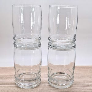 May include: Two clear glass tumblers stacked vertically in two pairs. Each glass features a smooth cylindrical body with a distinct horizontal ridge near the base and a slightly wider rim. The glass is transparent and colorless, showing a simple design.
