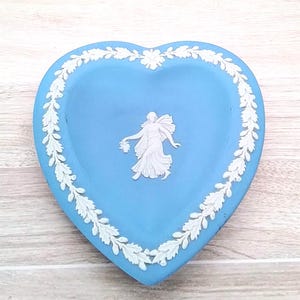 May include: A heart-shaped trinket dish in a light blue color with a white relief design. The dish features a white floral border and a central figure. A decorative piece for storing small items.