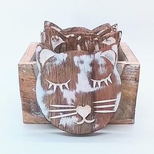May include: Set of six wooden cat-shaped coasters with a distressed, rustic finish. The coasters are brown and white, with closed eyes and heart-shaped noses. They are stored in a matching wooden holder, perfect for protecting surfaces from drink rings.