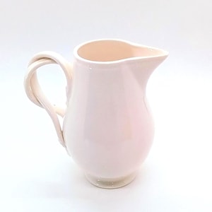 May include: A pale pink ceramic pitcher with a twisted handle and a spout for pouring. The pitcher has a rounded body and a smooth, glossy finish. The handle is a unique design element, adding visual interest to the piece.