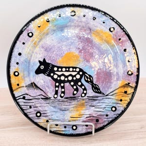 May include: A round ceramic plate featuring a black wolf illustration with white polka dots. The background is a painted wash of purple, blue, and yellow hues. The plate has a black rim decorated with small white dots and two mountain silhouettes.