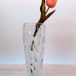 May include: A clear glass vase with a textured, swirling design, holding a single peach-colored tulip. The vase is tall and slender, with a wide opening. The tulip has green leaves and a brown stem.