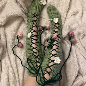 May include: A pair of sage green crochet socks featuring a lace-up design with dark green cord ties. The socks are decorated with small white crocheted flowers and dangling pink strawberry-shaped accents attached to the ends of the dark green cords.