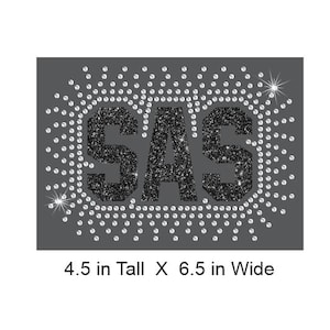 Puede incluir: Un diseño rectangular gris oscuro con las letras "SAS" en purpurina negra, rodeado por un círculo de pequeños diamantes de imitación transparentes. El diseño mide 11,4 cm de alto y 16,5 cm de ancho.