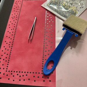 K&ouml;nnte beinhalten: Ein rotes Filztuch mit einem schwarzen gepunkteten Rand, eine Pinzette, ein blaues Applikationswerkzeug mit einem wei&szlig;en Pinsel und eine T&uuml;te mit klaren Strasssteinen.