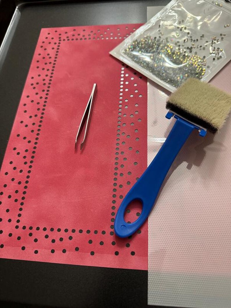 K&ouml;nnte beinhalten: Ein rotes Filztuch mit einem schwarzen gepunkteten Rand, eine Pinzette, ein blaues Applikationswerkzeug mit einem wei&szlig;en Pinsel und eine T&uuml;te mit klaren Strasssteinen.