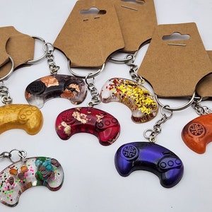 May include: A set of six colorful resin game controller keychains. Each keychain features a different color and design, including gold, red, black, orange, blue, and multi-colored. The keychains are attached to silver key rings.