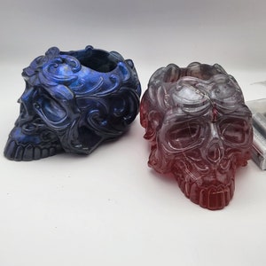 Puede incluir: Dos portavelas decorativos con forma de calavera, uno es azul con purpurina iridiscente y el otro es rojo con purpurina iridiscente.