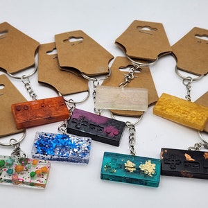 May include: A collection of colorful resin keychains in the shape of retro video game controllers. Each keychain features a different color and design, including glitter, gold leaf, and various embedded elements. They are attached to silver keyrings.