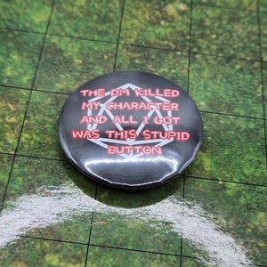 May include: A round, black button with the text "THE DM KILLED MY CHARACTER AND ALL I GOT WAS THIS STUPID BUTTON" in red and white. The button features a white geometric shape in the center.