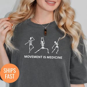 May include: Dark gray t-shirt featuring white skeleton figures in yoga poses. The text "MOVEMENT IS MEDICINE" is printed below the skeletons. The shirt has a round neck and short sleeves.