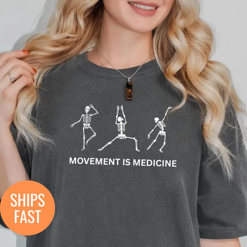 May include: Dark gray t-shirt featuring white skeleton figures in yoga poses. The text "MOVEMENT IS MEDICINE" is printed below the skeletons. The shirt has a round neck and short sleeves.