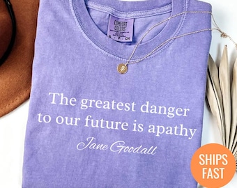May include: Violet Comfort Colors t-shirt with the quote "The greatest danger to our future is apathy" by Jane Goodall in white text. A gold necklace with a pendant rests on the shirt. The shirt is folded.