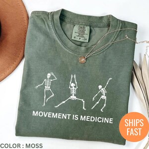 May include: Moss green t-shirt with a white skeleton graphic in yoga poses. The text "MOVEMENT IS MEDICINE" is printed below the graphic. A gold necklace is draped on the shirt. The shirt is labeled "COMFORT COLORS".