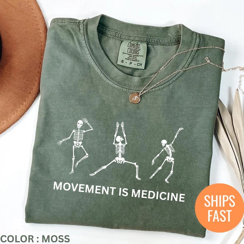 May include: Moss green t-shirt with a white skeleton graphic in yoga poses. The text "MOVEMENT IS MEDICINE" is printed below the graphic. A gold necklace is draped on the shirt. The shirt is labeled "COMFORT COLORS".