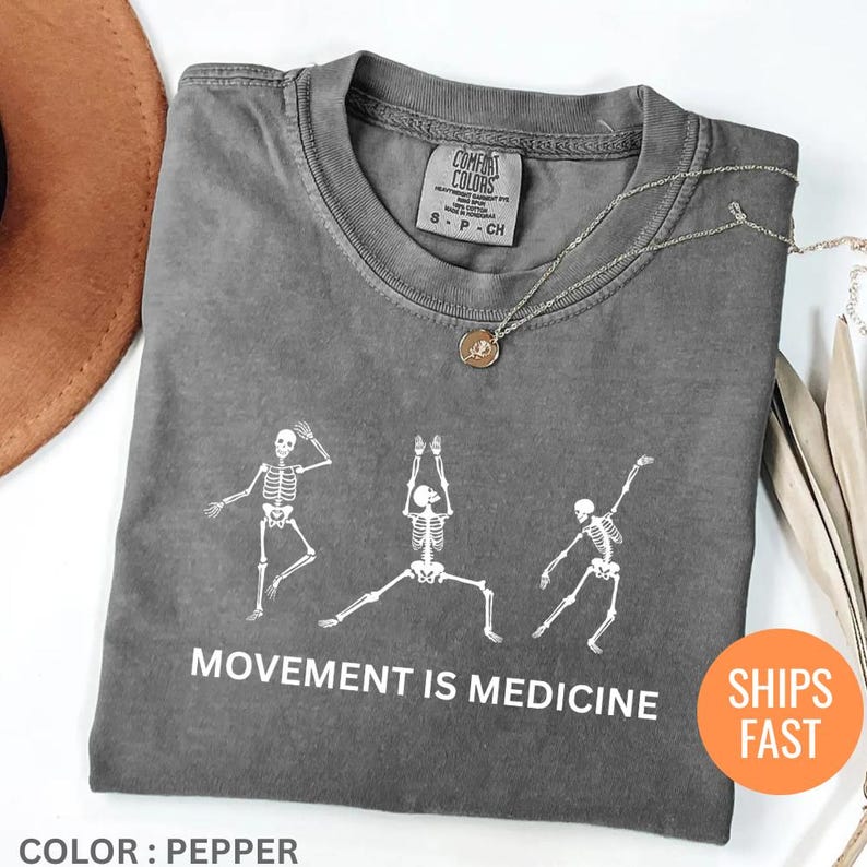 May include: Pepper-colored t-shirt with a white graphic of three skeleton figures in yoga poses. The text "MOVEMENT IS MEDICINE" is printed below the skeletons. A gold necklace is draped across the shirt. The shirt is labeled "COMFORT COLORS".