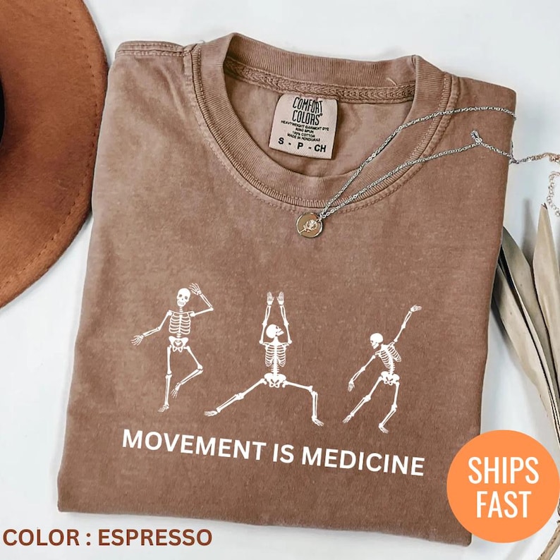 May include: Espresso-colored t-shirt with a white skeleton graphic in various yoga poses. The text "MOVEMENT IS MEDICINE" is printed below the skeletons. A silver necklace with a pendant is draped on the shirt.