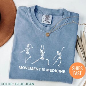 May include: Blue jean colored t-shirt with a white skeleton graphic in various yoga poses. The text "MOVEMENT IS MEDICINE" is printed below the skeletons. A gold necklace is draped across the shirt.