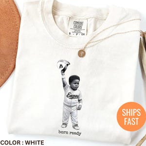 Peut inclure: T-shirt blanc avec une illustration en noir et blanc d'un jeune joueur de baseball levant une casquette. Le maillot porte l'inscription "Expos" et "born ready" en bas. Un collier doré est posé sur le t-shirt.