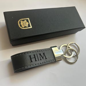 Könnte beinhalten: Ein schwarzer Lederschlüsselanhänger mit der Gravur H|M, einem silberfarbenen Metallbeschlag und zwei runden Schlüsselringen. Der Schlüsselanhänger wird mit einer rechteckigen schwarzen Geschenkbox geliefert, die ein goldenes Emblem mit dem Text ECHTES LEDER zeigt.