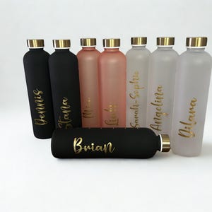 May include: A collection of personalized water bottles in various colors, including black, pink, and frosted. Each bottle has a gold lid and is inscribed with a name in gold script. The bottles are arranged on a white surface.