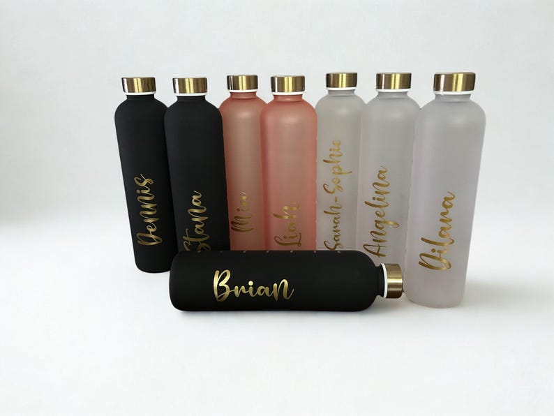 May include: A collection of personalized water bottles in various colors, including black, pink, and frosted. Each bottle has a gold lid and is inscribed with a name in gold script. The bottles are arranged on a white surface.
