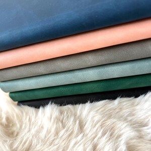 May include: A stack of colorful leather sheets in shades of blue, peach, gray, and green. The leather is stacked on a fluffy, white, faux fur surface. The colors are vibrant and the texture of the leather is visible.