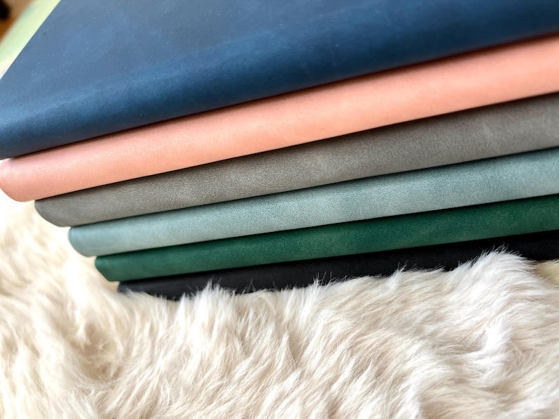 May include: A stack of colorful leather sheets in shades of blue, peach, gray, and green. The leather is stacked on a fluffy, white, faux fur surface. The colors are vibrant and the texture of the leather is visible.