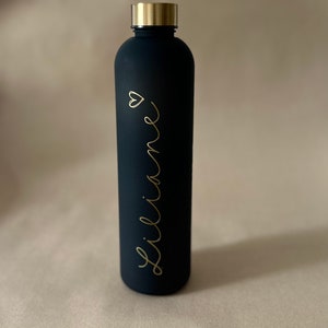 May include: A black water bottle with a gold-colored lid. The bottle has the name "Liliane" in gold script with a small heart symbol. The bottle is cylindrical and appears to be made of metal.