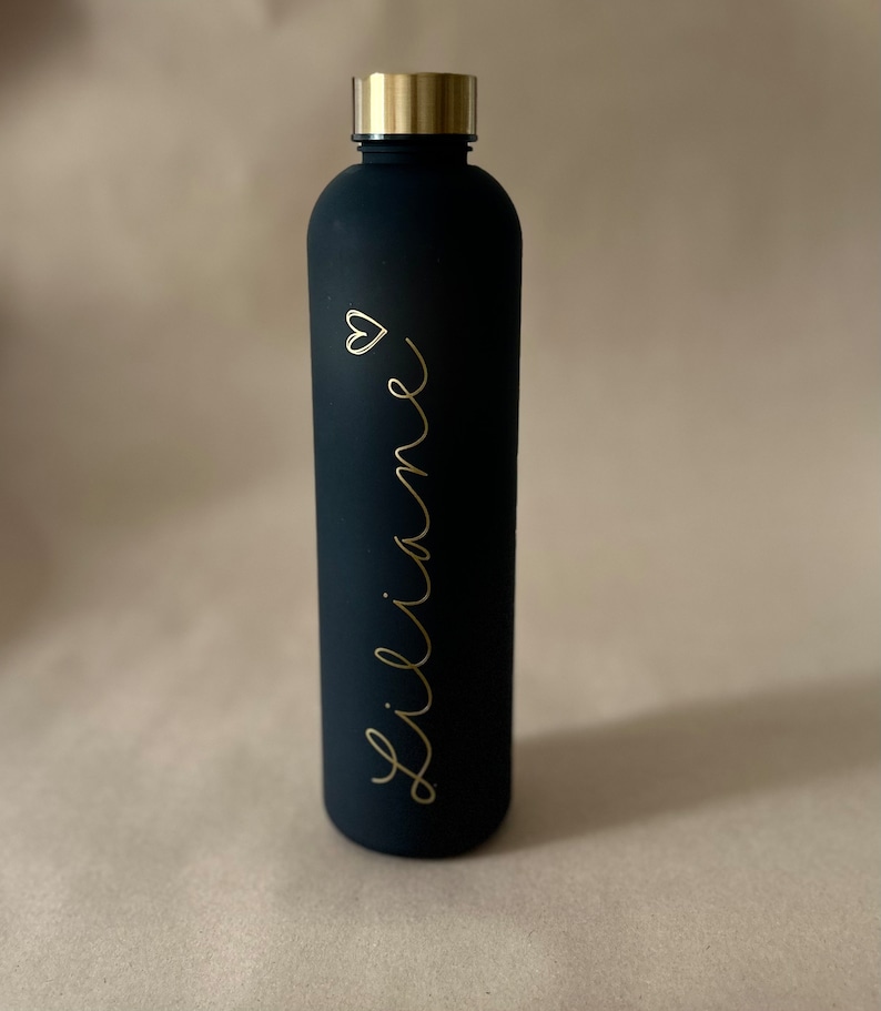 May include: A black water bottle with a gold-colored lid. The bottle has the name "Liliane" in gold script with a small heart symbol. The bottle is cylindrical and appears to be made of metal.
