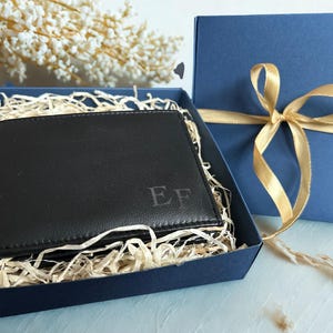 May include: A black bi-fold wallet featuring the embossed letters Ef on the front corner. The wallet is nestled inside a dark blue gift box filled with light-colored paper filler, accompanied by a matching blue lid tied with a gold ribbon.