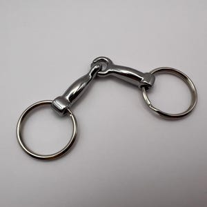 Könnte beinhalten: Ein poliertes Metallpferdegebiss mit zwei großen kreisförmigen Ringen und einem gebogenen Mittelgelenk. Das Gebiss ist dafür konzipiert, in das Maul eines Pferdes gelegt und zur Kontrolle an den Zügeln befestigt zu werden. Das Metall hat eine glänzende, reflektierende Oberfläche.