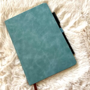 May include: A teal-colored journal with a textured cover and a brown ribbon bookmark. A black pen with a blue band is tucked inside the journal. The journal is resting on a fluffy, off-white surface.